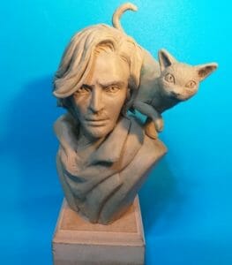 Picture of a clay sculpted bust of Caleb, with Frumpkin on his shoulder. The sculpture is placed on a plinth in front of a blue background. Caleb has medium length hair, a beard and is wearing a jacket with a large collar. Frumpkin stands on his shoulder, brushing against his hair with his tail raised.