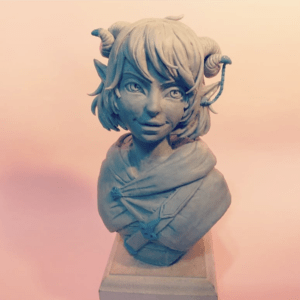 Picture of a clay sculpted bust of Jester placed on a plinth in front of a pink background. She has ram-like horns with a chain running from one to her pointed ear. She has short hair with bangs. She is wearing a hooded cloak with a strap around her shoulder.