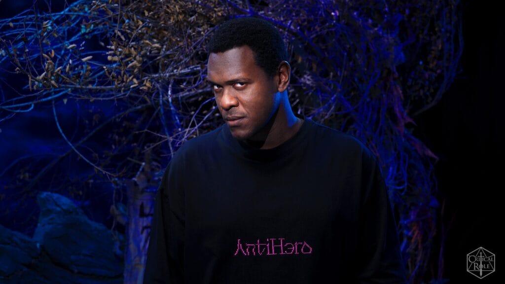 Portrait of a man with short black hair under blue neon lighting, wearing a black sweatshirt with pink 'AntiHero' text