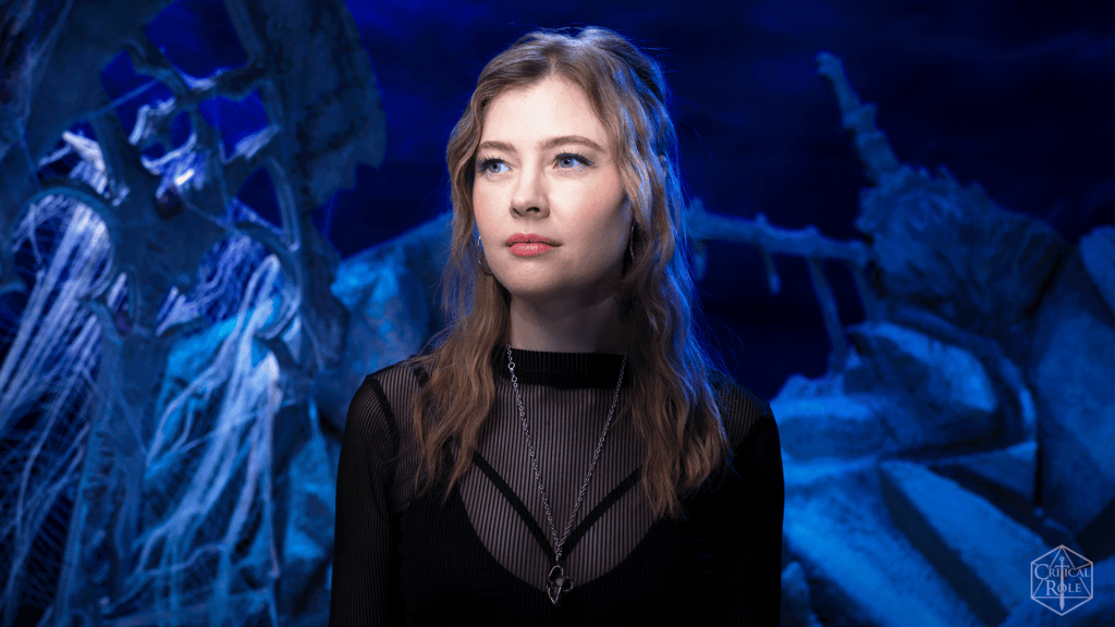 Portrait of a young woman with long wavy hair in a black mesh top and silver necklace, standing in a blue-lit cave with webs and jagged rocks.