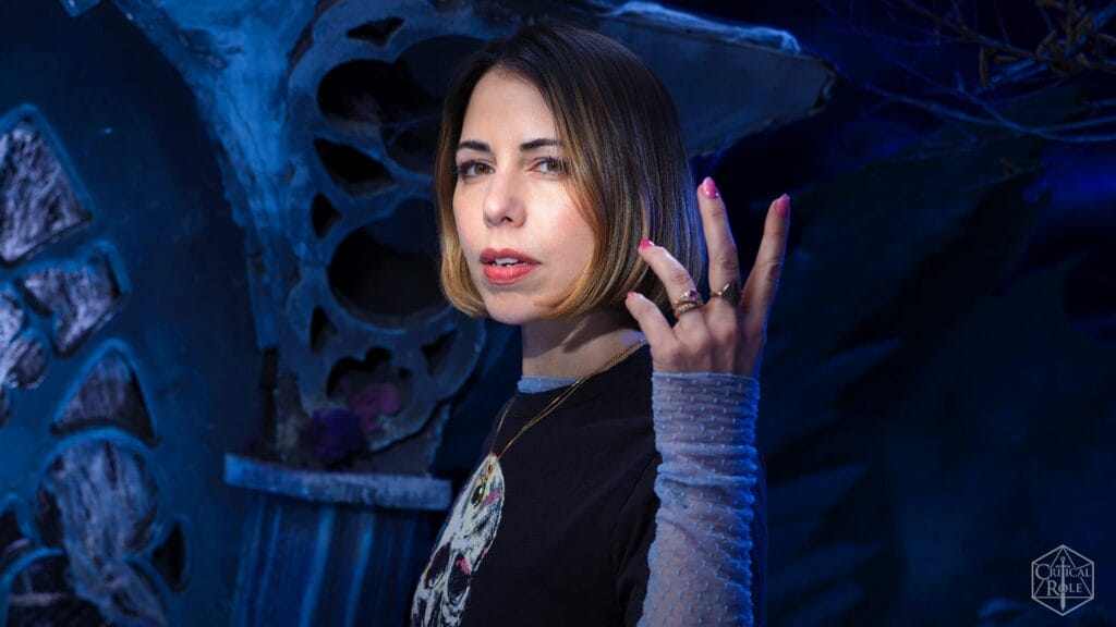 Portrait of a woman with shoulder-length brown hair under blue lighting, wearing a black shirt and multiple rings, against a large gear-like backdrope.