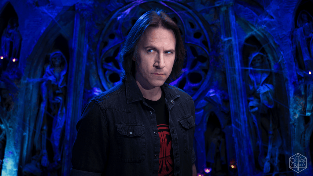 Man with shoulder-length hair wearing a dark denim jacket stands in a blue-lit fantasy cavern filled with statues in the background.