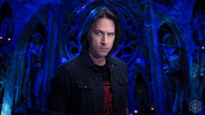 Man with shoulder-length hair wearing a dark denim jacket stands in a blue-lit fantasy cavern filled with statues in the background.