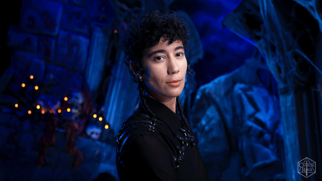 Portrait of a person with short curly hair and braids in a blue-lit cavern, wearing a black outfit with metal studs in the background.