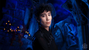 Portrait of a person with short curly hair and braids in a blue-lit cavern, wearing a black outfit with metal studs in the background.
