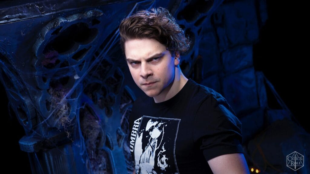 Portrait of a man with dark hair wearing a black graphic t-shirt, in a moody blue-lit, web-filled background.