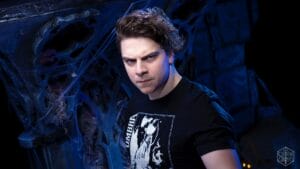 Portrait of a man with dark hair wearing a black graphic t-shirt, in a moody blue-lit, web-filled background.